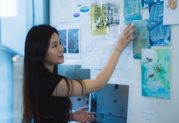 The woman is identified as Hazel Yuhang Zhang, an artist touching a painting. The photo shows her in an indoor setting, possibly a studio, with a focus on her face and the artwork she is working on.