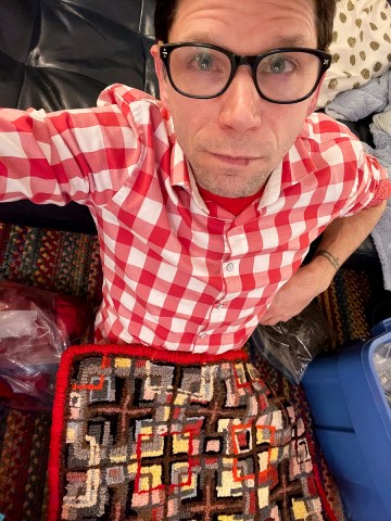 The image is a self-portrait of Jamie Girourard wearing glasses. The person is indoors, wearing a red shirt and the photo focuses on their face. The image also includes a tartan or plaid pattern.