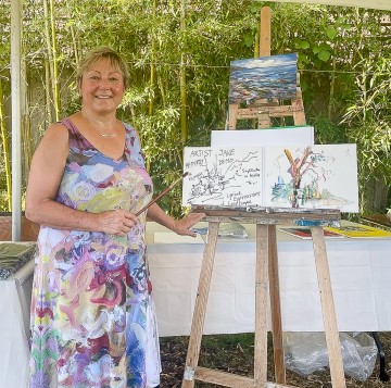 A woman stands beside an easel displaying a painting. The artwork is part of the workshop 'Colour Movements in Painting: Abstracting Ambleside'.