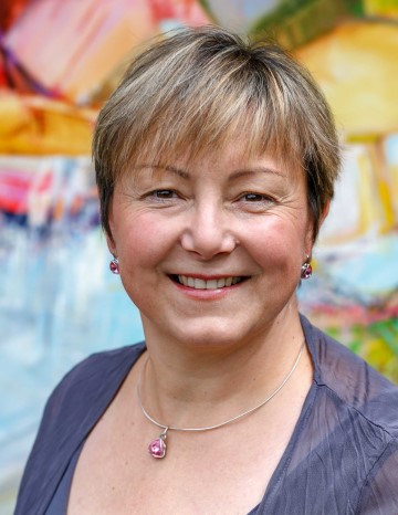 Jane Appleby, expressive painter, smiling in front of a painting with short hair, posing for a photo.