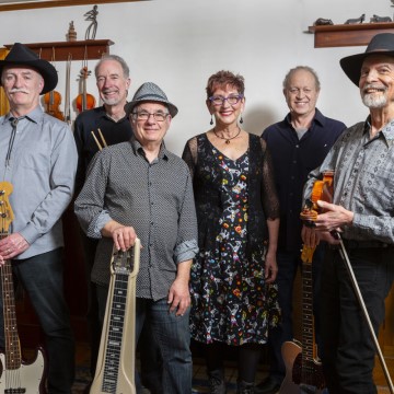 The image shows a group of people holding musical instruments, including guitars. They are wearing various types of hats and appear to be posing indoors against a wall. The group is likely associated with the bands SUE MALCOLM & THE SWINGIN' DOORS.
