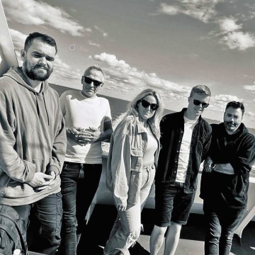 The photo features a group of people posing for a picture on a coast, with a clear sky and clouds in the background. They are wearing casual clothing, including jeans, and some are smiling. The image is in black and white and includes a mix of men and women.