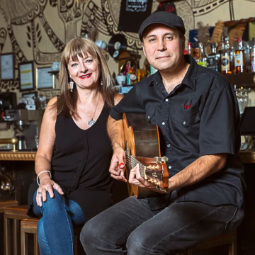 The image features a man and a woman posing for a picture. The man is wearing a black hat and holding a guitar. The woman is wearing makeup and both individuals seem to be in a pub or bar setting.