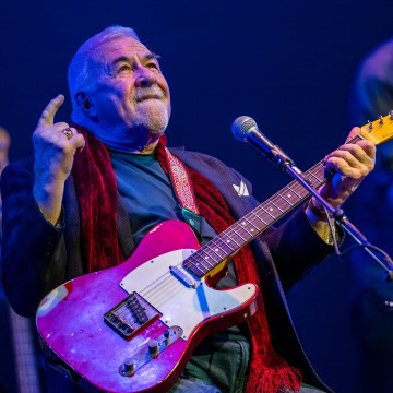 The image shows a man playing a guitar at a concert. He is a musician named Jim Byrnes and is performing with an electric guitar. The setting includes a microphone and purple lighting.