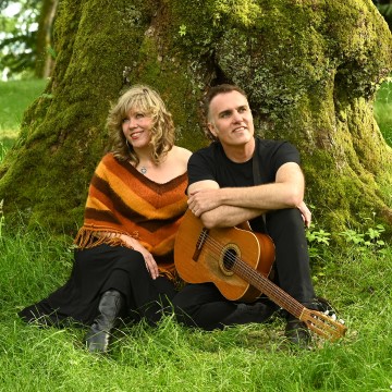 The image features a man and a woman sitting on grass with a guitar, both of them smiling. The setting is outdoors, possibly under a tree. The man is holding the guitar while the woman is sitting beside him. The overall atmosphere appears to be relaxed and enjoyable.