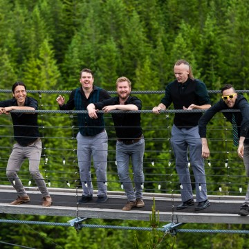 The photo shows a group of men from The Whiskeydicks band standing  on a bridge outdoors. They are wearing casual clothing and the surrounding area includes grass and trees.
