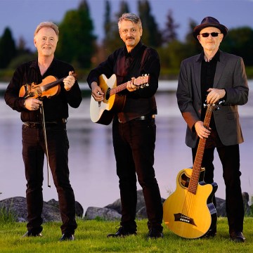 The image is of a group of men from the Tiller's Folly band holding guitars and mandolins while standing outdoors on grass. The men are wearing casual clothing and footwear.