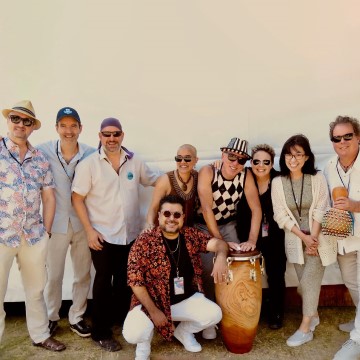 The image depicts a group of people from the Timba Cartel band posing for a photo indoors. They are standing against a wall, with some holding musical instruments like drums. The group includes men and women, all smiling for the photo.