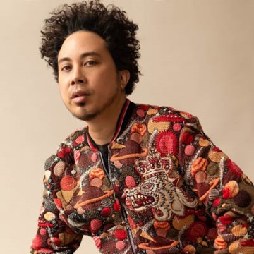 The image shows a person with curly hair, identified as Warren Dean Flandez, a singer. He is indoors, wearing a red robe, standing in front of a wall.