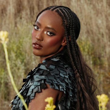The image features a person with long hair, specifically Zada Singer, in an outdoor setting. The person is wearing braids and is surrounded by grass. The photo focuses on the human face and fashion style of the individual.