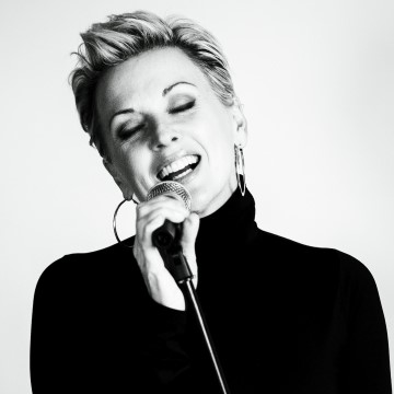 The photo is of a woman singing into a microphone. It is in black and white and shows the woman indoors against a wall. The woman has a smile on his face.