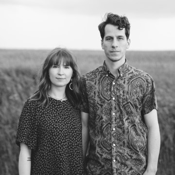 The image features a man and woman posing for a picture in an outdoor setting. The man and woman are both smiling. The photo is in black and white and includes elements like the sky, grass, and field. They are dressed in casual clothing. The context of the image is related to the Anna & Regan Luth band.