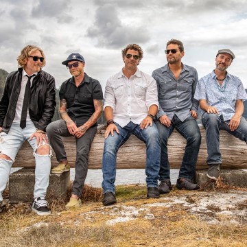 The image features a group of men sitting on a rock. They are outdoors, surrounded by grass and under a cloudy sky. The men are wearing casual clothing like jeans and jackets. The men appear to be from the band Buddy & The Scarecrow.