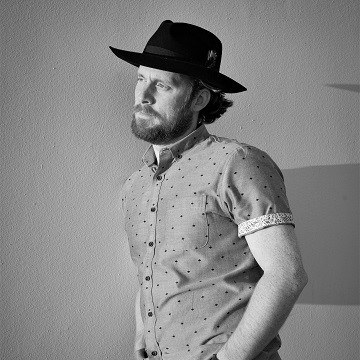 The image features a man wearing a hat indoors. He is standing and the photo is in black and white. The man is identified as Ivan Hartle, a singer.