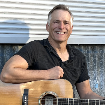 The image shows Jeff Standfield, a singer, sitting with a guitar. He is smiling while holding the acoustic guitar.