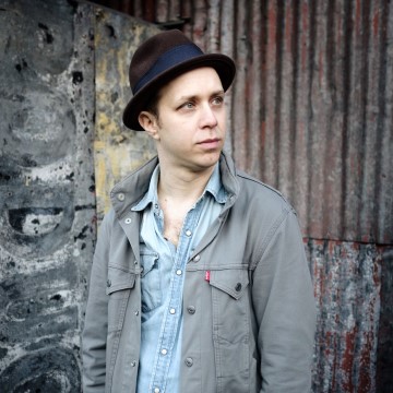 The photo is of a person wearing a hat and is related to Jesse Waldman, a singer. The person in the image is a man dressed in outerwear, a coat, a cap, and a shirt, standing outdoors on the street.