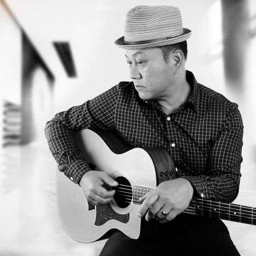 The image shows a person playing a guitar indoors. They are wearing a fedora and a shirt. The photo is in black and white. The person is identified as Jim Kwan Duo.