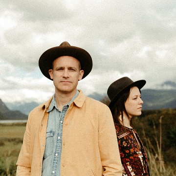 The image features a man and a woman wearing hats and smiling. They are standing in a field with mountains in the background. The man is wearing a cowboy hat and the woman is wearing a fedora. It seems like they are posing for a picture.