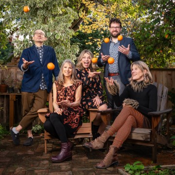 The image shows a group of people posing for a photo outdoors, with a background of trees and a bench. The group includes women, girls, and possibly family members. They are smiling and dressed in casual clothing. Additionally, there is a mention of a band playing with oranges in the additional context provided.