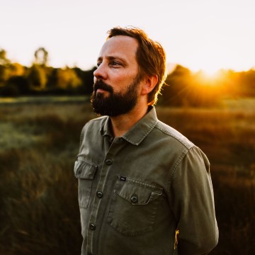 The image shows a man with a beard, identified as Ryan McMahon, a singer. He is standing outdoors during sunset, with the sky in the background. The man has facial hair and a moustache.