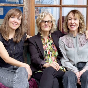 The photo depicts the band "The Luckies," consisting of Shari Ulrich, Jeanne Tolmie, and Hilary Grist, sitting on a bench. The women are smiling and wearing casual clothing.