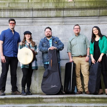 The image shows a group of people, including Pat Chessell, standing on stairs. They are all smiling and holding musical instruments. The setting is outdoors. The group is casually dressed in jeans and trousers, and some are carrying luggage and bags.