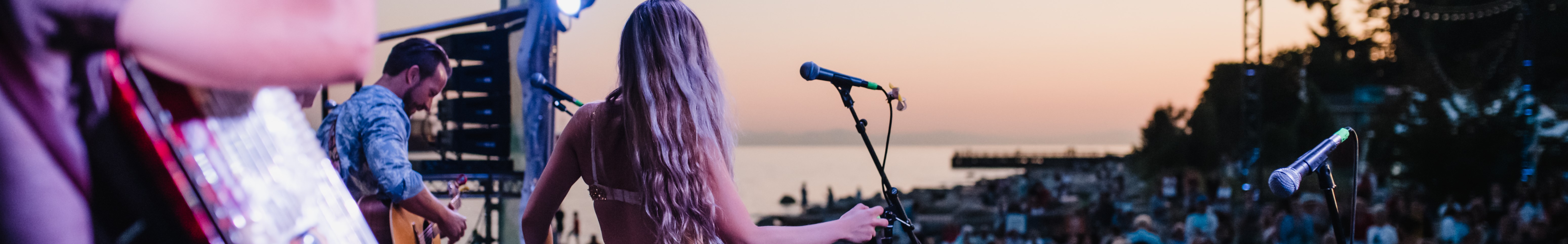 Silhouetted figure holding a microphone on a music stage at sunset. Female singer and guitarist performing in the background.