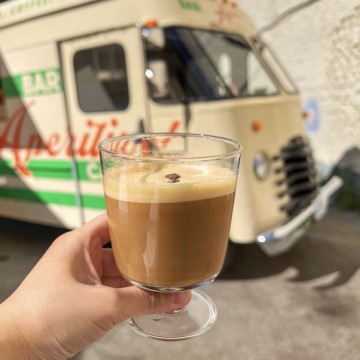 The image shows a hand holding a glass with coffee latte. The setting is at Aperitivo Coffee Truck. Tags for the image include beverage, coffee, food, drinkware, caffeine, cup, coffee milk, person, outdoor, and drink.