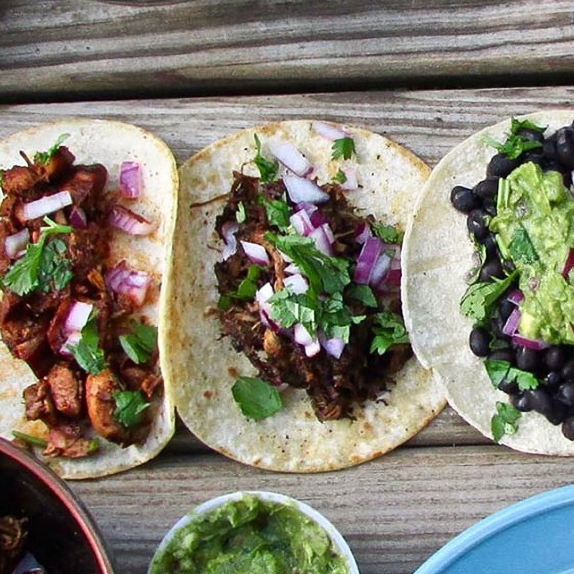 The image shows a group of tacos on a table from Dos Amigos food truck. The tacos seem to be a variety of Mexican and Tex-Mex food, including Korean taco, gringas, chalupa, and tostada. Ingredients like guacamole, leaf vegetables, and corn tortillas are also included.