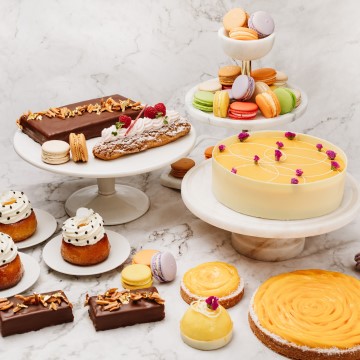 The image is of a table full of cakes, showcasing a variety of desserts and baked goods such as cakes, cupcakes, and pastries. The display includes items like buttercream cakes, petit fours, and cream-filled treats. The spread is decorated with icing and food coloring, creating a visually appealing array of sweet treats.