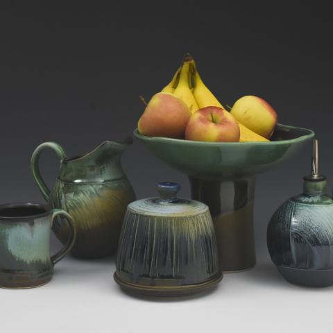 The image is a still life photograph featuring a bowl of fruit and a pitcher. The items are placed on a table and include ceramic pottery and earthenware. The setting appears to be indoors and the image may be related to Duncan Tweed Ceramics.