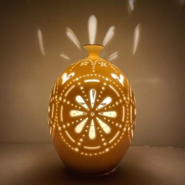 The image shows a lit up lantern created by Dogi Jagi Ceramics. The photo features a gold-colored still life photography of the illuminated vase against a wall with amber lighting.