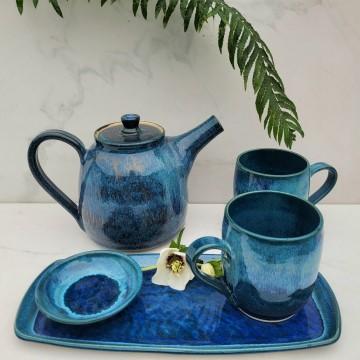 The image features a teapot and teacups placed on a blue plate. The items are ceramic and part of Mathew Freed Pottery. The setting includes indoor plants and kitchenware.
