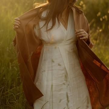 The image is of a woman with long hair in an outdoor setting, possibly in a field. She is wearing a dress and standing on grass. The image is associated with Nomi Designs.