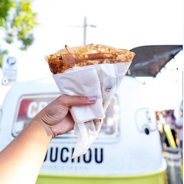 The image shows a hand holding a crepe, specifically a "chou chou crepe." The background includes the sky and the setting appears to be outdoors