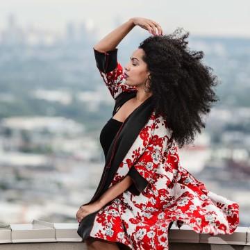 The image shows a woman posing for a picture, likely during a photoshoot for KOME Clothing. She is outdoors and is wearing a dress, showcasing fashion. The focus is on the woman's face and clothing.